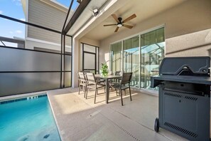 Um pátio externo com piscina, mesa e cadeiras, ventilador de teto e churrasqueira.