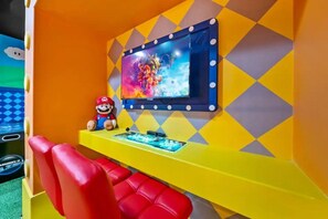 Um espaço de jogos com um boneco do Mario, uma tela de TV exibindo um jogo e uma escrivaninha amarela com uma cadeira vermelha.