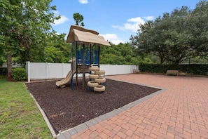 Ein Spielplatz mit Rutsche und Kletterbereich, umgeben von einem Pflasterweg und Grünflächen.