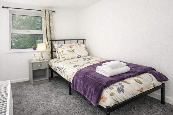 A single bed with a floral duvet, a bedside table with a lamp, and a window with curtains.
