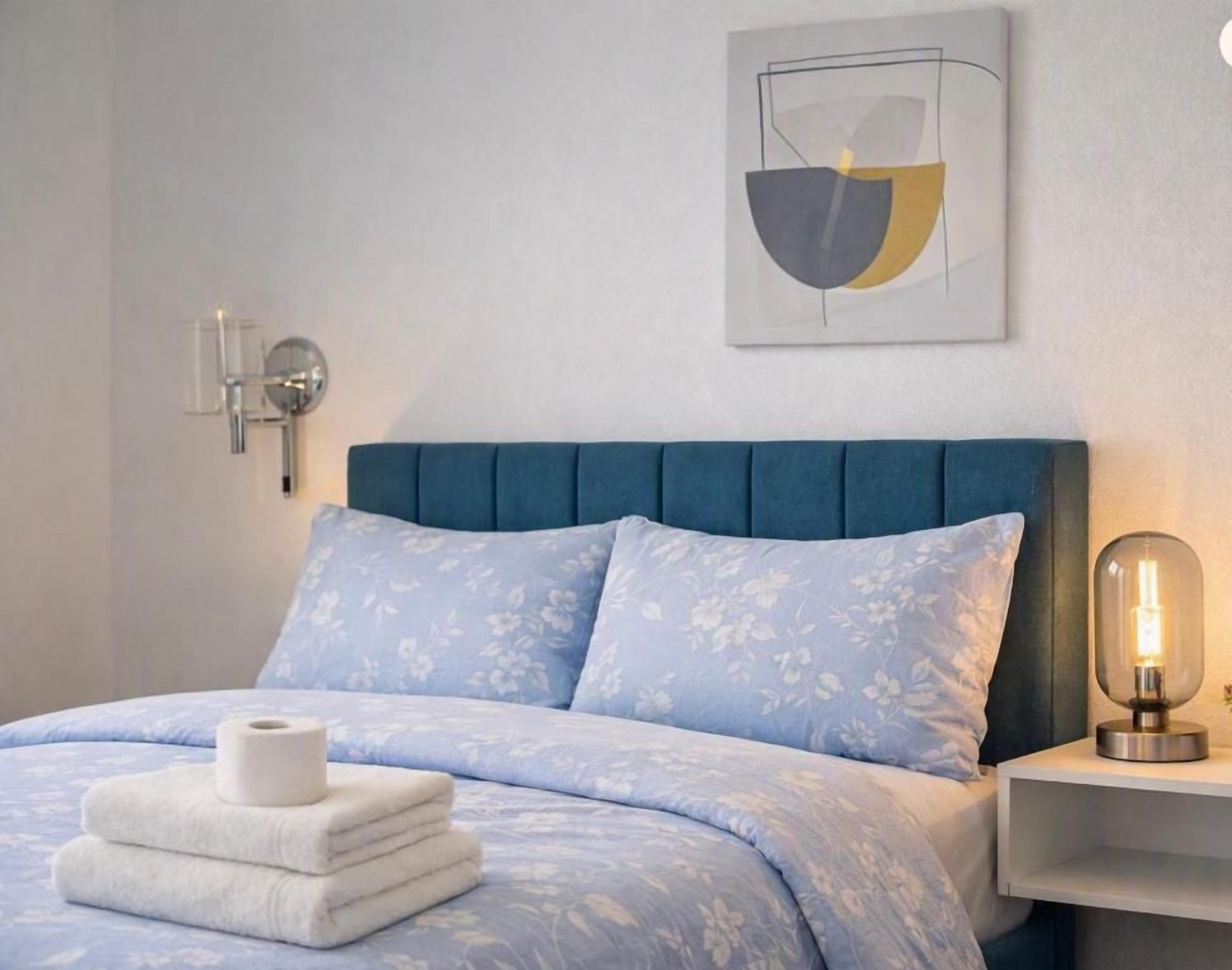 A bedroom with a blue upholstered headboard, a bed with a floral patterned duvet, a bedside table with a lamp, and a wall-mounted artwork.
