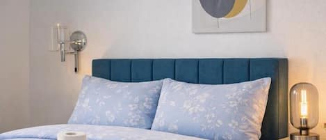 A bedroom with a blue upholstered headboard, a bed with a floral patterned duvet, a bedside table with a lamp, and a wall-mounted artwork.