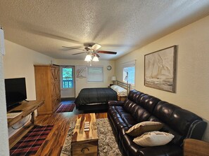 A room with a ceiling fan, a bed, a leather sofa, a television, a wooden cabinet, and a painting on the wall.
