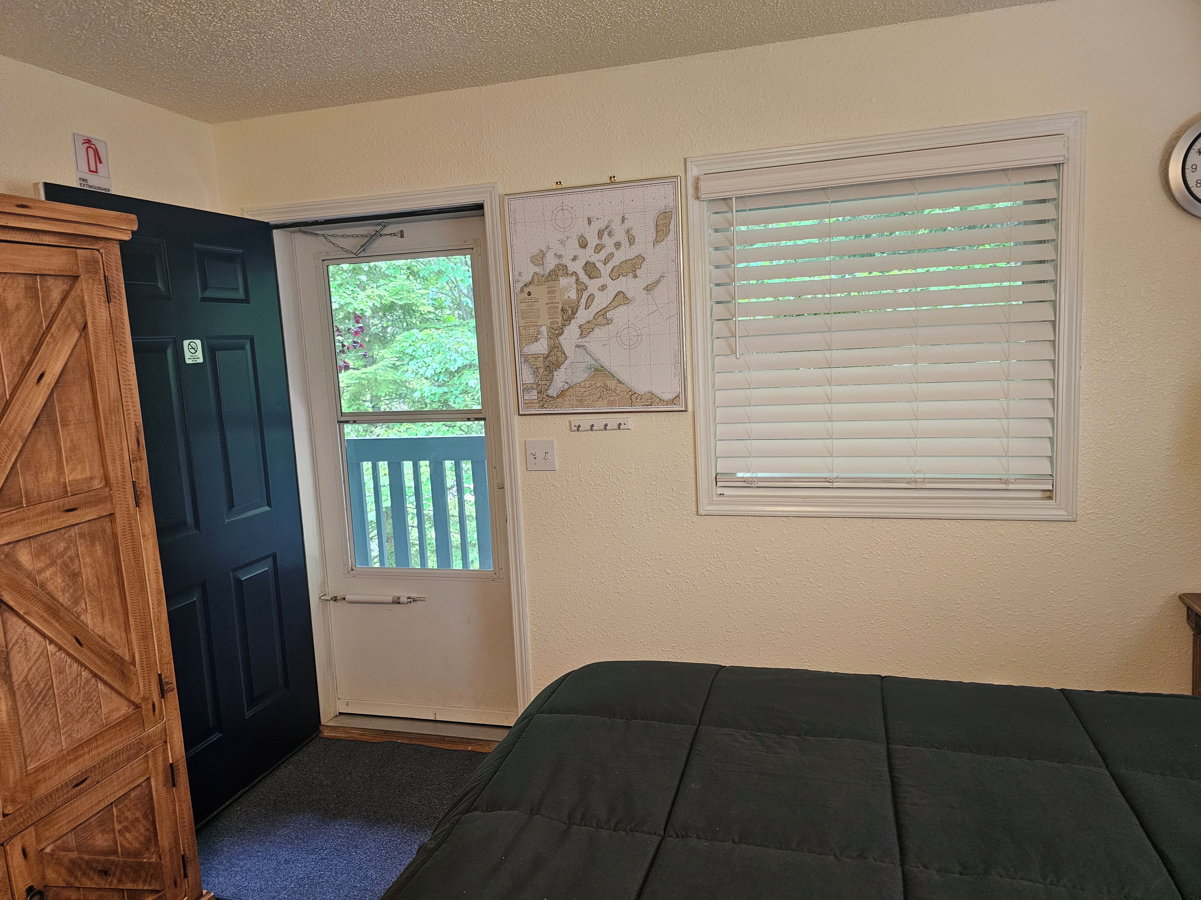A room with a door, a window with blinds, a bed, and a wall-mounted clock.