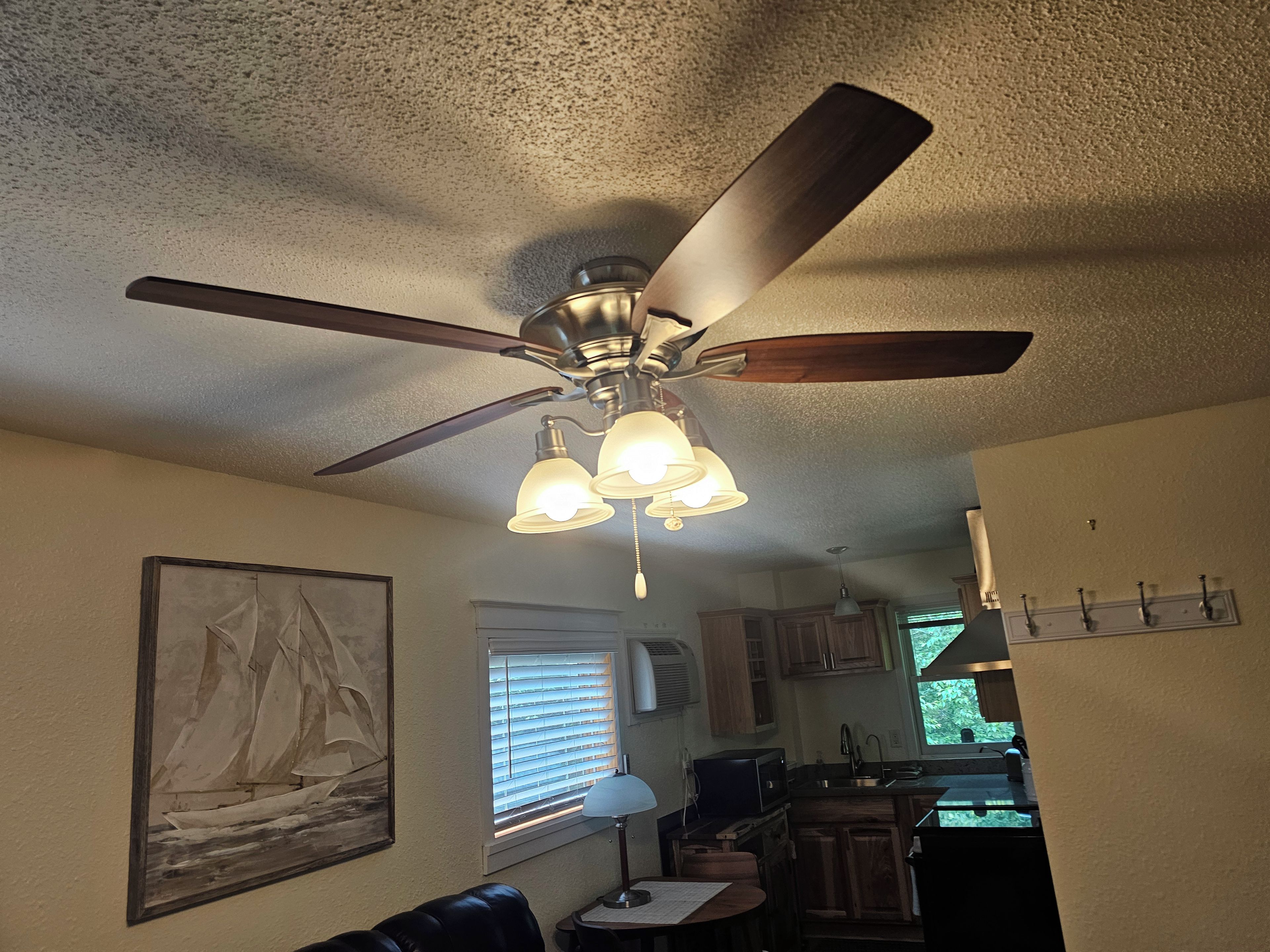 A ceiling fan with lights, a sailboat painting, a window with blinds, and a kitchen area.