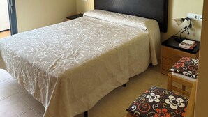 Ein Schlafzimmer mit einem Bett, einem Nachttisch, einem Sessel, einem Schreibtisch mit Lampe, einer Deckenleuchte und einer Wandsteckdose.