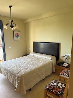Ein Schlafzimmer mit einem Bett, einem Nachttisch, einem Sessel, einem Schreibtisch mit Lampe, einer Deckenleuchte und einer Wandsteckdose.