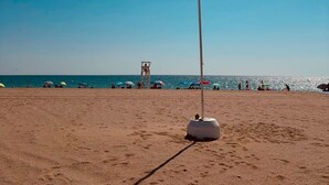 Ein Strand mit einer Fahnenmast, einem Rettungsschwimmturm und Sonnenschirmen im Hintergrund.