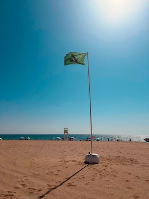 Ein Strand mit einer Fahnenmast, einem Rettungsschwimmturm und Sonnenschirmen im Hintergrund.