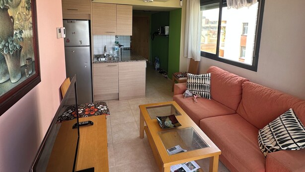 Ein Wohnzimmer mit Sofa, Couchtisch und Fernseher. Im Hintergrund ist eine Küche mit Kühlschrank und Mikrowelle zu sehen.