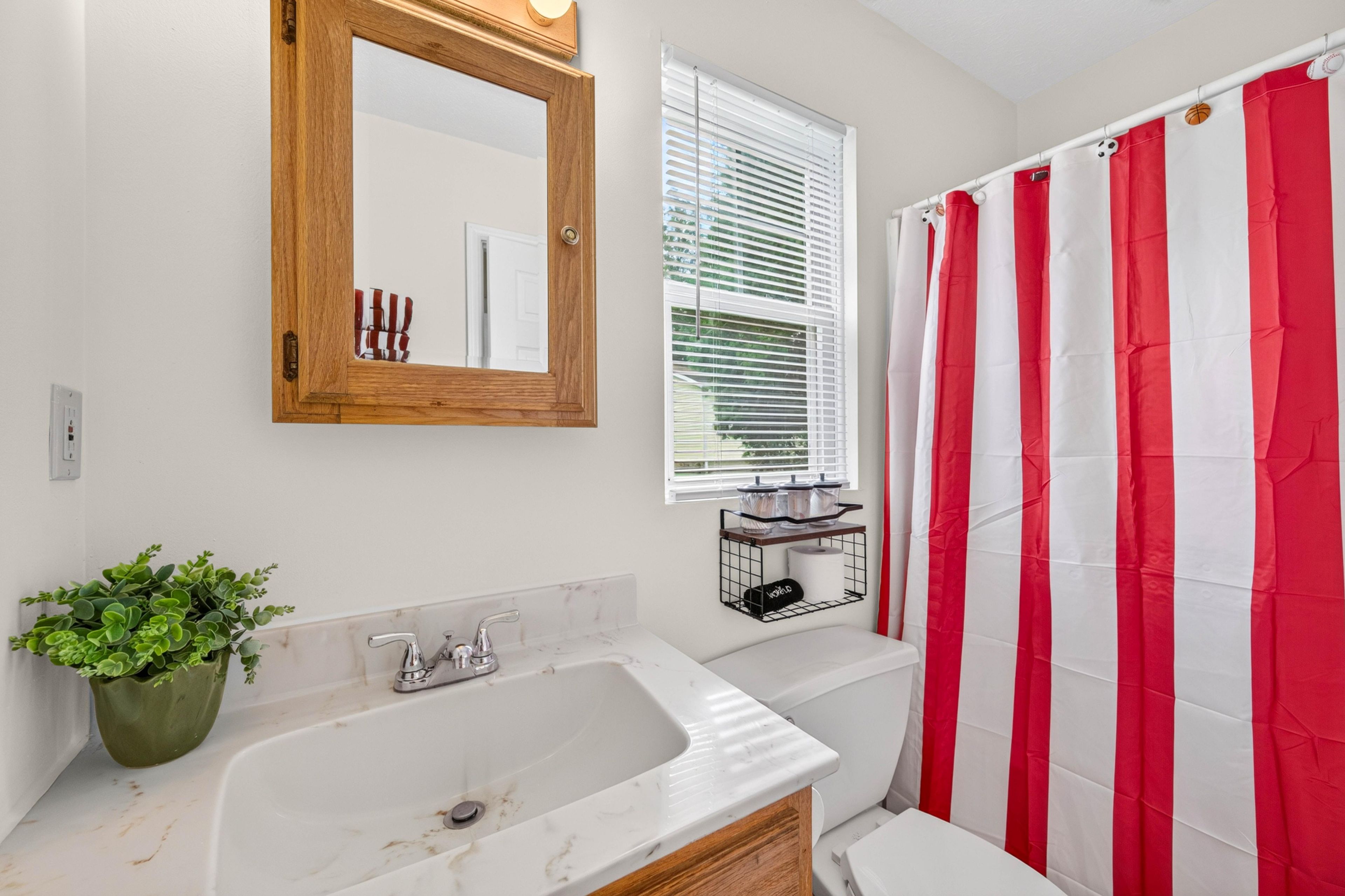Une salle de bain avec un lavabo blanc, un meuble en bois, un miroir, une fenêtre avec des stores et un rideau de douche à rayures rouges et blanches.