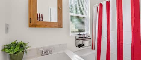 Une salle de bain avec un lavabo blanc, un meuble en bois, un miroir, une fenêtre avec des stores et un rideau de douche à rayures rouges et blanches.