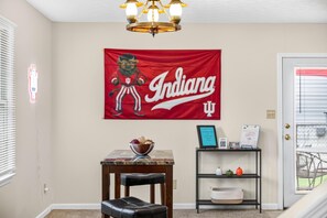 A room with a flag, a table with a bowl, a chair, and a shelf with decorative items. - Hoosier House on S. Park Ave. Newly renovated, Close to IU Campus, (Bloomington)