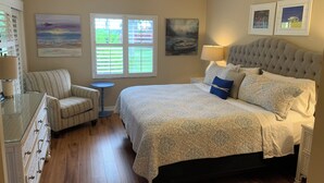 Quarto com uma cama grande, uma cadeira, uma mesinha e obras de arte nas paredes.