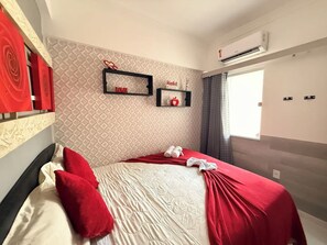 A hotel room with a bed, red pillows, a wall-mounted air conditioner, and a window with curtains.