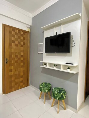 A room with a wooden door, a flat-screen TV mounted on a white wall, and two green stools with wooden legs. - 204 - Cozy 2-bedroom apartment. (Belém)