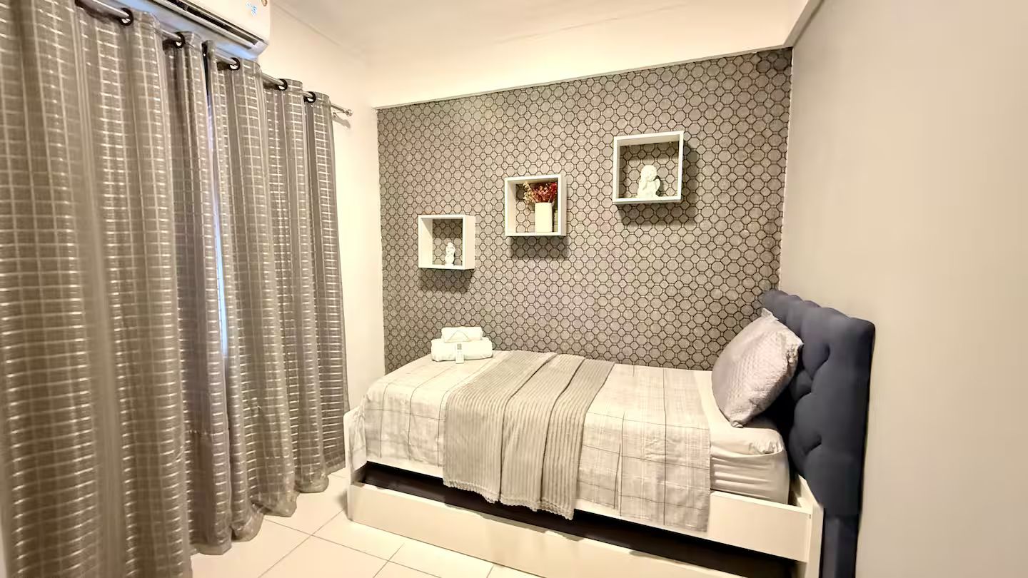 Quarto com cama, cortinas, cabeceira e prateleiras na parede com objetos decorativos.