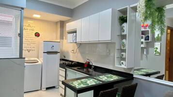 Uma cozinha compacta com ilha central, armários brancos e um pequeno espaço de estar com duas cadeiras.