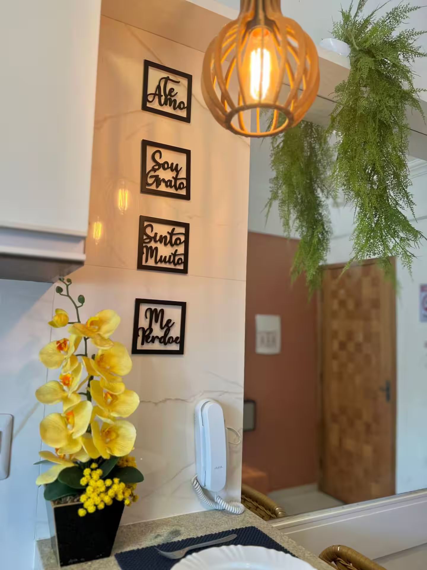 A bathroom with a mirror, a wall-mounted light fixture, a potted plant, and decorative wall art with text.