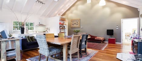 Een ruime loft met een hoge plafond, een eethoek met een houten tafel en rieten stoelen, een woonkamer met een rode bank, en een keuken met witte kasten.