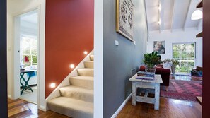 Eine Treppe mit beigem Teppich, einer roten Wand und einem Holzboden. Ein kleiner Tisch mit Büchern und einer Pflanze steht daneben, sowie ein großes gerahmtes Kunstwerk an der Wand.