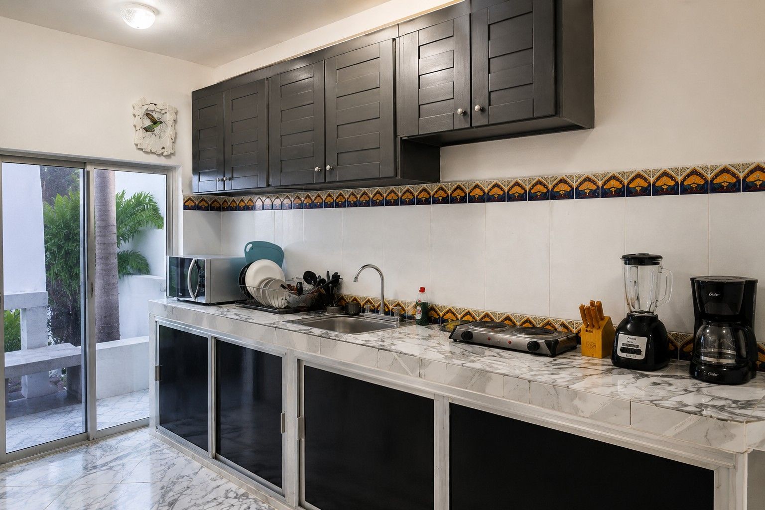 Una cocina con armarios oscuros, salpicadero de azulejos, un fregadero, una estufa, una licuadora y una cafetera.