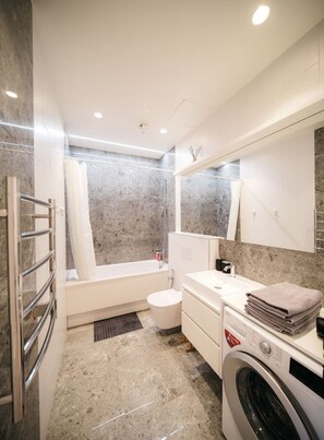 A modern bathroom with a bathtub, toilet, and washing machine. - Modern 1-Bedroom Apartment on Dzintaru Prospekts (Jūrmala)