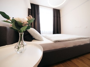 A modern bedroom with a bed, a bedside table with a vase of flowers, and a window with curtains. - Modern 1-Bedroom Apartment on Dzintaru Prospekts (Jūrmala)