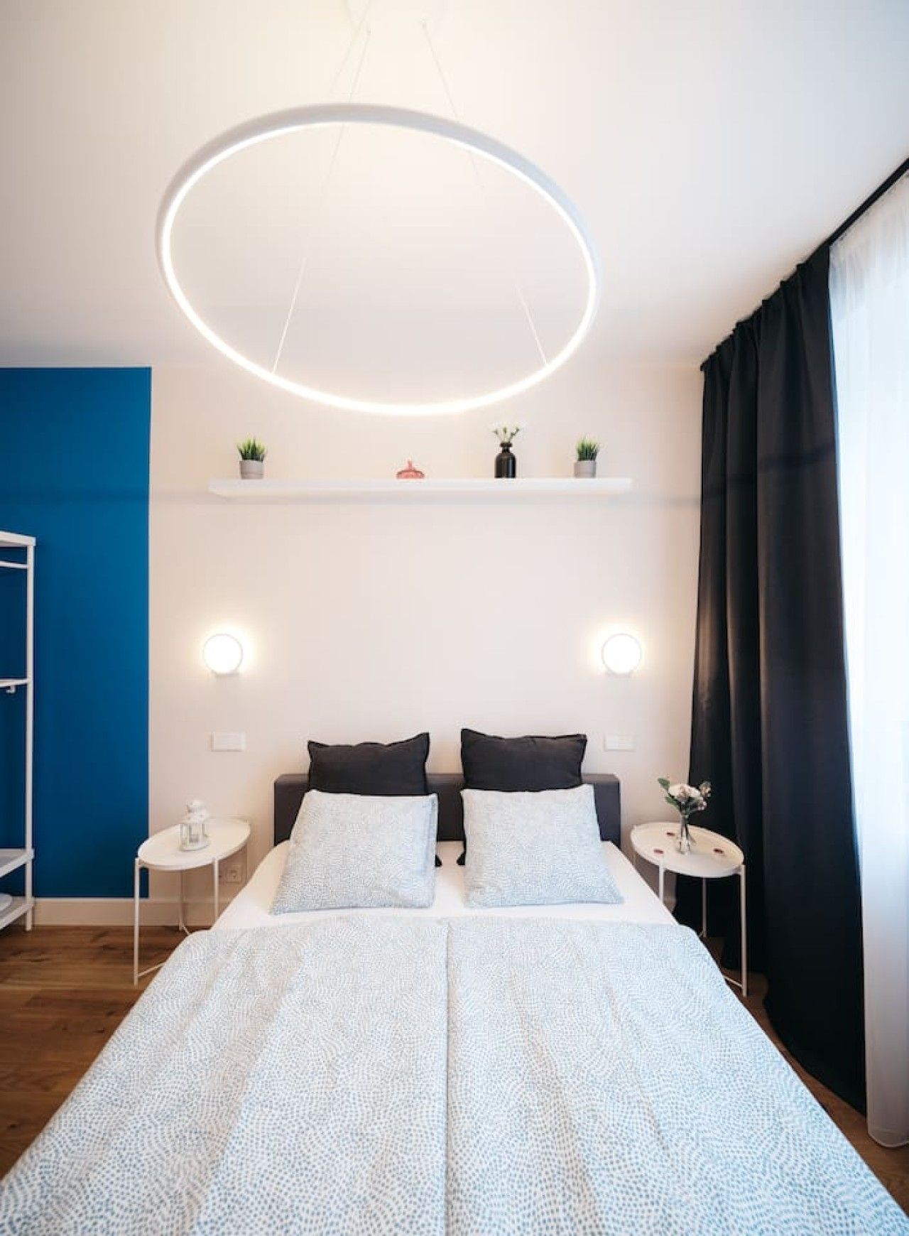 A bedroom with a bed, bedside tables, a hanging light fixture, a blue accent wall, and a window with curtains.