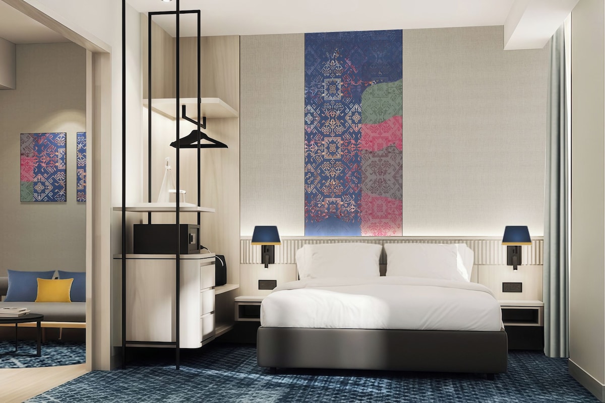 Ett modernt hotellrum med en säng, sängbord, ett skrivbord, en stol och en stor, abstrakt målning på väggen.