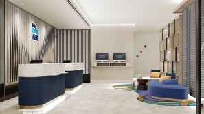 En hotellobby med modern design, med en receptionsdisk, två bildskärmar och en sittgrupp med en soffa och ett soffbord.