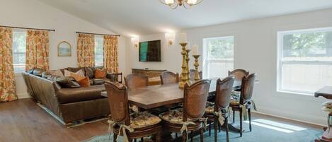 A dining area with a wooden table and chairs, a sofa, a chandelier, and a large window.