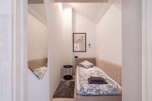 Ein schmaler Flur führt zu einem Schlafzimmer mit einem Bett, einem kleinen Nachttisch, einem gerahmten Bild an der Wand und einem Spiegel.