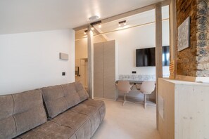 A modern living room with a sofa, a dining area with chairs, a TV mounted on a brick wall, and a wooden cabinet. - Stylish 2-Bedroom Attic Loft with AC & Workspaces (Rīga)