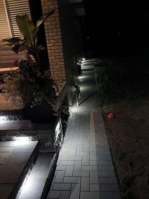 Un sendero nocturno que conduce a un edificio con fachada de ladrillo y una planta en maceta a la izquierda.