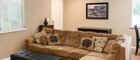 A living room with a sectional sofa, ottoman, and a painting on the wall.