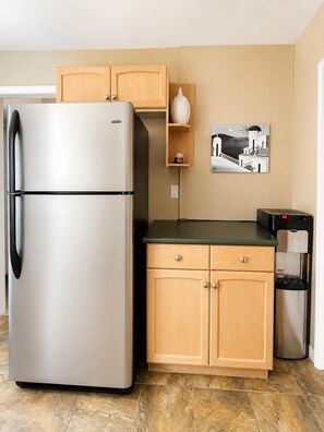 A compact kitchen with a stainless steel refrigerator, wooden cabinets, a small shelf with a decorative item, and a water dispenser.