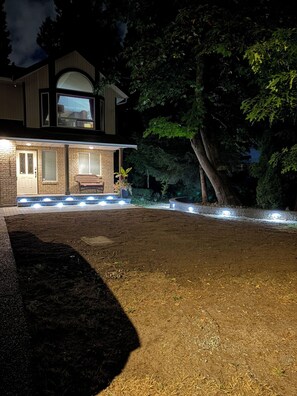 Una casa de dos pisos con fachada de ladrillo, un porche con banca y un camino de entrada bien iluminado.