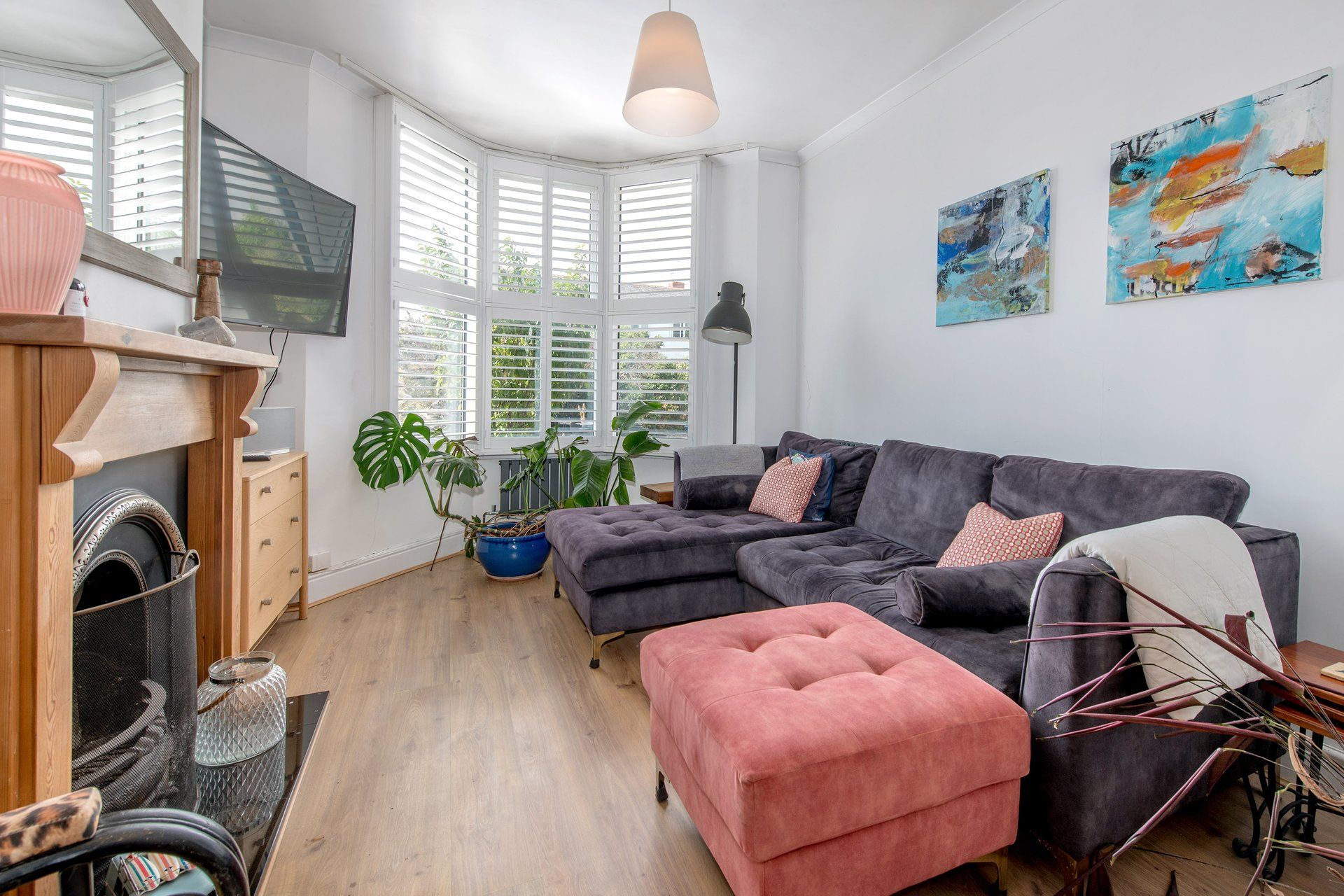 A modern living room with a sectional sofa, a pink ottoman, and a fireplace. There are two large windows with shutters, a ceiling light, and two abstract paintings on the wall.