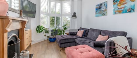 Ein modernes Wohnzimmer mit einer Eckcouch, einem pinkfarbenen Hocker und einem Kamin. Zwei große Fenster mit Rollläden, eine Deckenleuchte und zwei abstrakte Gemälde an der Wand sind zu sehen.