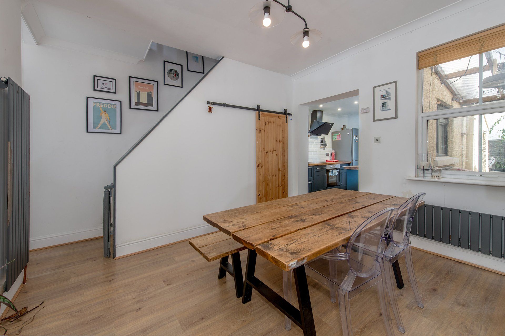 A dining area with a wooden table and clear plastic chairs, a staircase, and a kitchen visible through an open door.