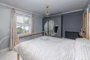 Quarto com cama, espelho redondo, radiador e janela com cortinas.