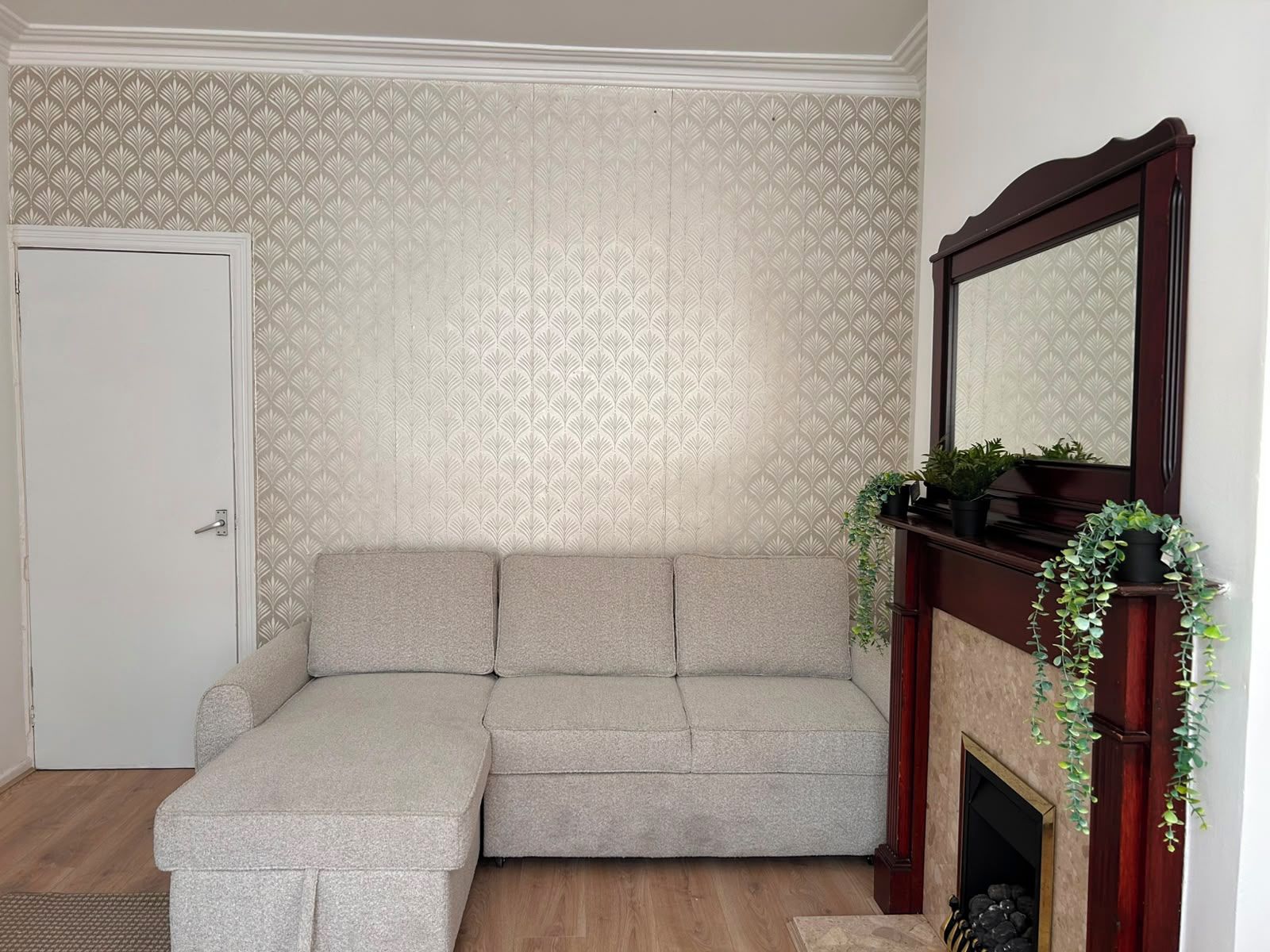 A corner room with a sectional sofa, a fireplace with a mirror, and a patterned wallpaper.