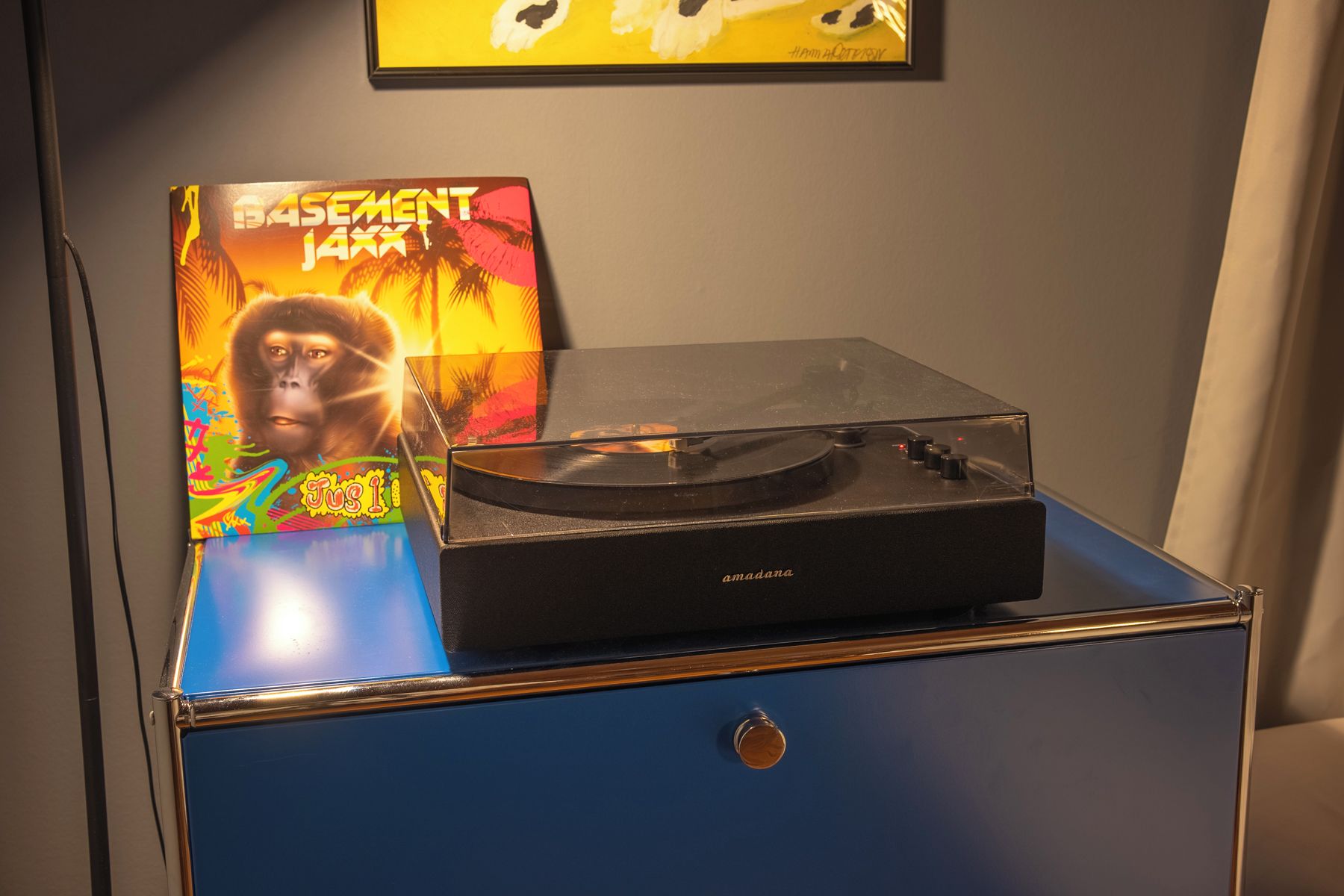 A turntable on a blue cabinet with a record sleeve labeled 'Basement Jazz' and a painting above it.