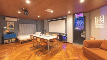 A hotel room with a bed, a dining table with chairs, a TV screen, a dartboard, and a wall-mounted air conditioning unit.
