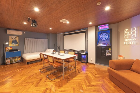 A hotel room with a bed, a dining table with chairs, a TV screen, a dartboard, and a wall-mounted air conditioning unit.