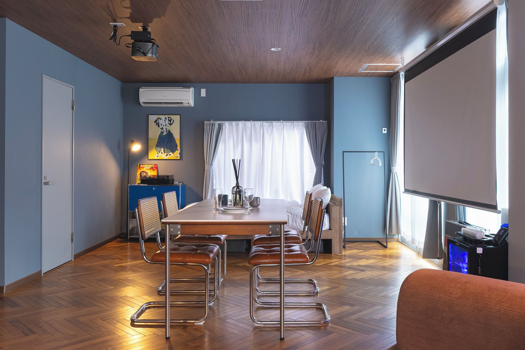 Un salón de conferencias con una mesa de madera, sillas de metal, una pantalla de proyector y un amplio ventanal con cortinas.