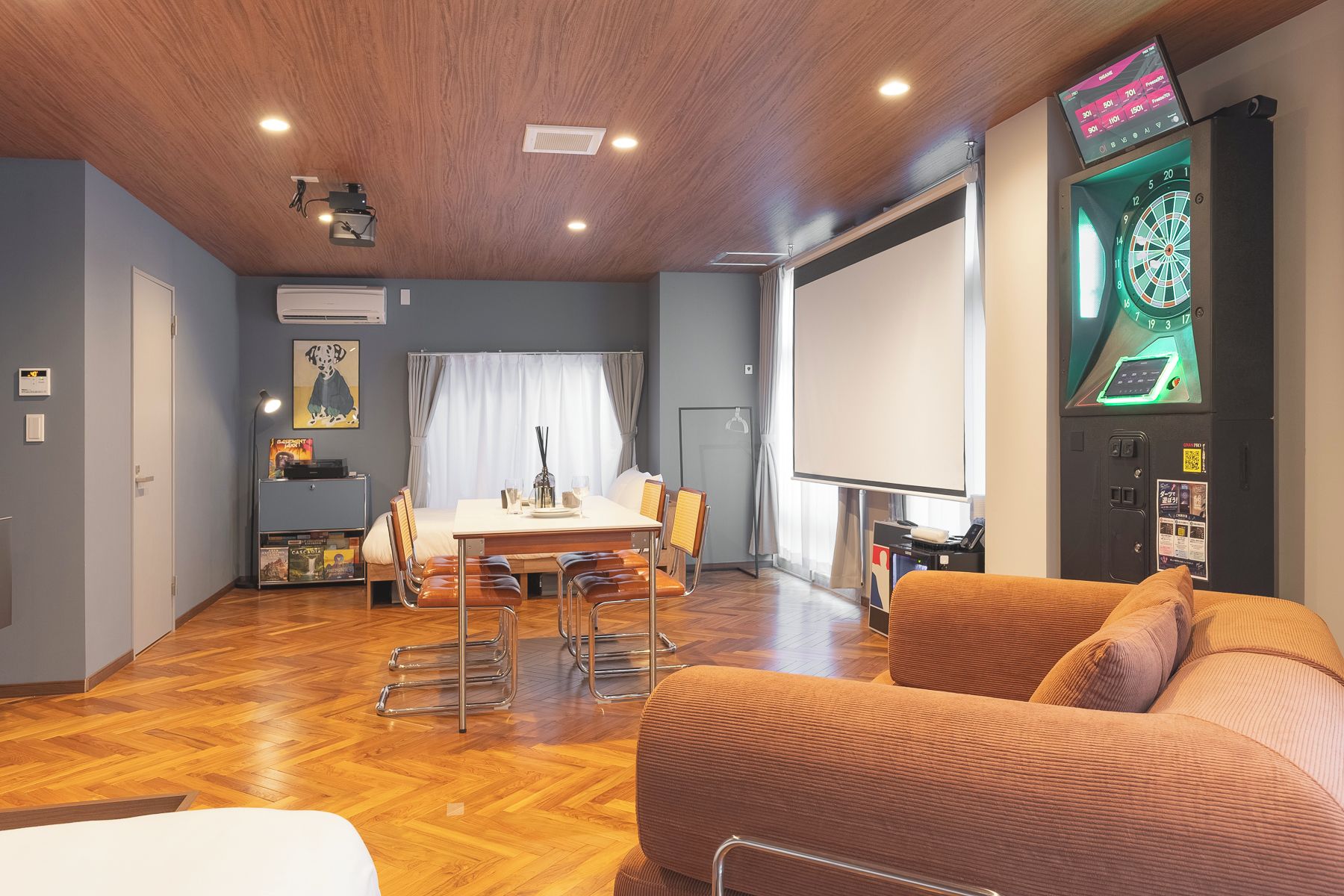 A room with a wooden ceiling, a dining table with chairs, a couch, a dartboard, and a projector screen.