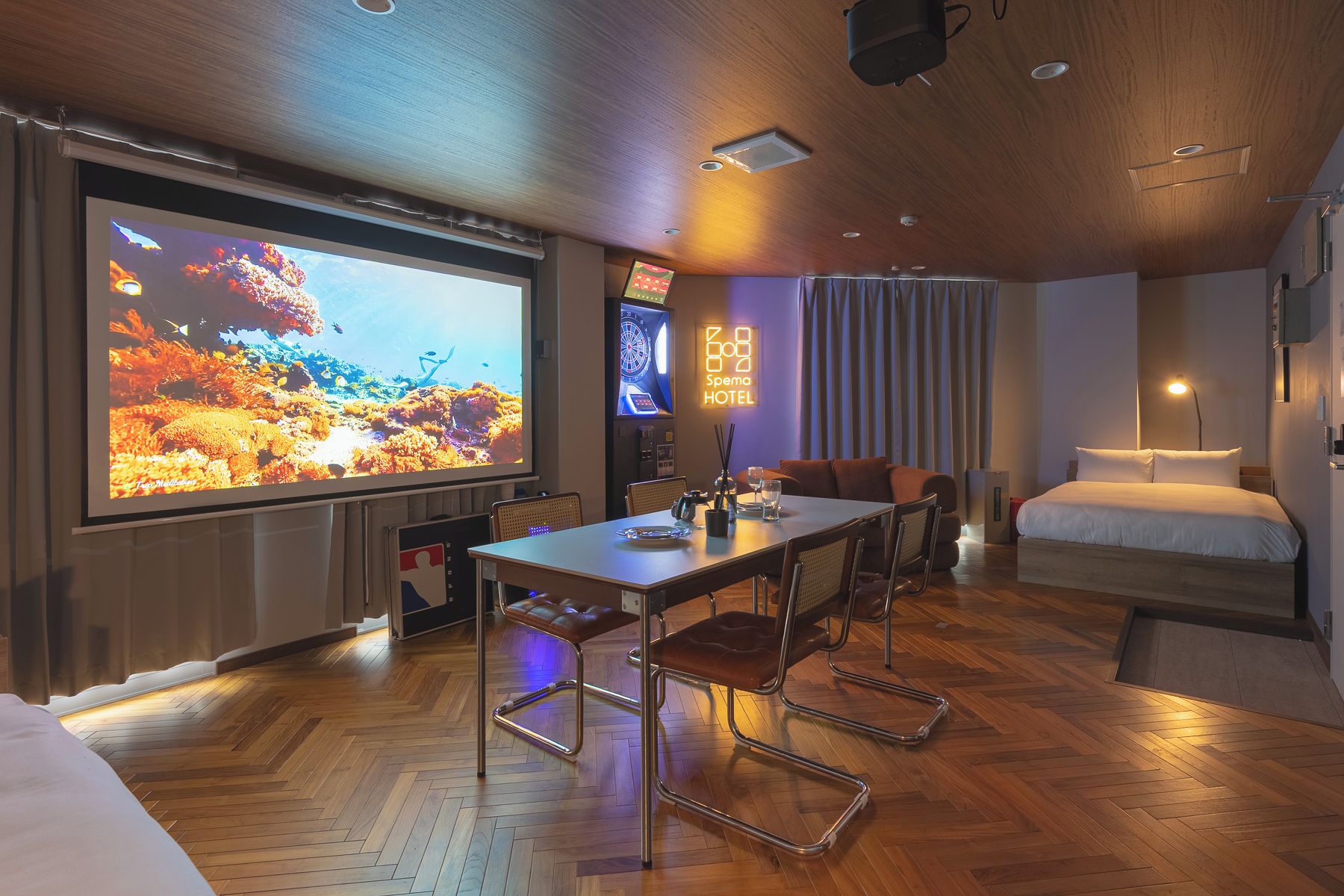 Habitación de hotel con una pantalla grande que muestra una escena submarina, un comedor con mesa y sillas, una cama y un televisor montado en la pared.