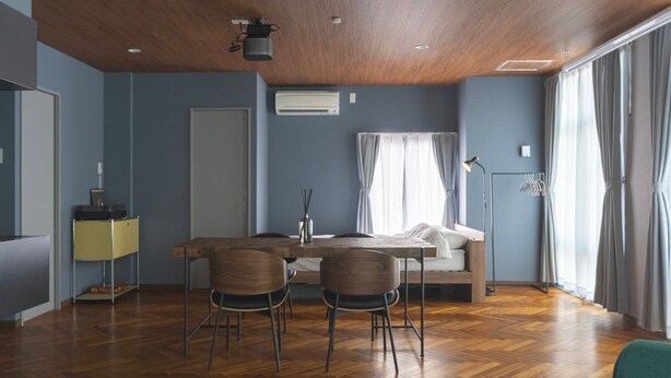 A modern room with a wooden ceiling, a bed, a desk with chairs, a standing lamp, and a large window with curtains.